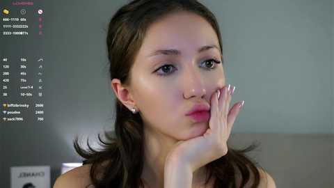 Video of a young woman with light skin, long dark hair, and light makeup, resting her chin on her hand, looking contemplative. Background shows a blurred room with a calendar and clock visible.