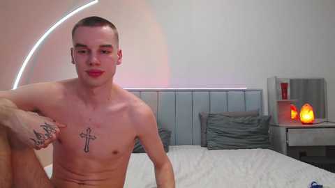 Video of a young, shirtless, fair-skinned man with short hair and a tattoo of a cross on his chest, sitting on a bed with gray pillows and a white sheet in a modern, minimalist bedroom.
