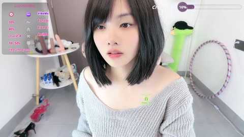 Video of a young East Asian woman with straight black hair and fair skin, wearing a grey off-shoulder sweater, in a cluttered room with a white table, shoes, and a hula hoop.
