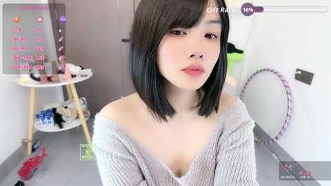 A video of an East Asian woman with straight black hair and fair skin, wearing a grey off-shoulder sweater, posing indoors. The background includes a white wall and a small table with scattered items.