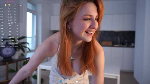 Video of a smiling, fair-skinned woman with long, auburn hair, wearing a white lace camisole, standing in a modern kitchen with white cabinets and dark countertops.