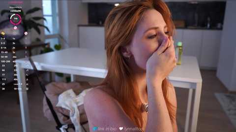 Video of a fair-skinned, red-haired woman in a white top, sitting at a white table, covering her mouth, with a man's hand on her shoulder in a modern kitchen.