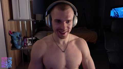 Video of a shirtless, smiling, muscular, white man with a short haircut wearing large headphones, in a dimly lit bedroom with a TV showing a blue screen.
