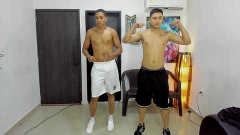 Video of two shirtless, muscular young men flexing in a sparsely decorated room with a dark wooden door, a chair, and colorful artwork on the wall.