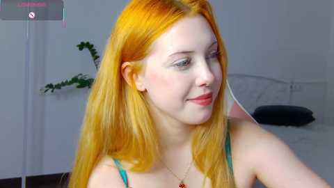 Video of a young Caucasian woman with long, vibrant orange hair, fair skin, and blue eyes, wearing a teal tank top, smiling in a bedroom with a white bed, green plant, and a dark pillow.