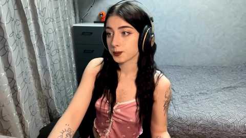 Video of a young woman with long black hair, wearing headphones, a pink satin top, and tattoos, sitting in a bedroom with gray curtains and a bed with a geometric patterned sheet.