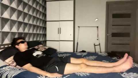 Video of a young man with dark hair and sunglasses lying on a bed with a blue patterned comforter, wearing a black t-shirt and shorts, in a modern, geometric-patterned bedroom.