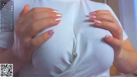A video of a person with large breasts, wearing a white t-shirt, grasping the fabric to reveal cleavage. The background is blurred, showcasing a QR code in the bottom left corner.