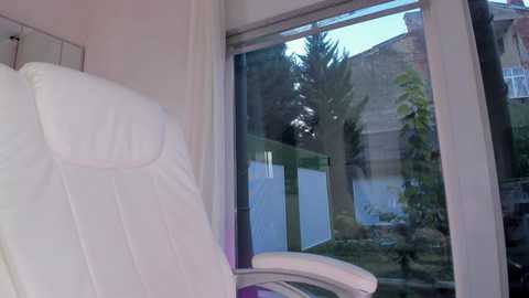 A video of a modern, white recliner chair next to a large window, showing a green garden with trees and a brick wall outside, with soft natural light filtering in.