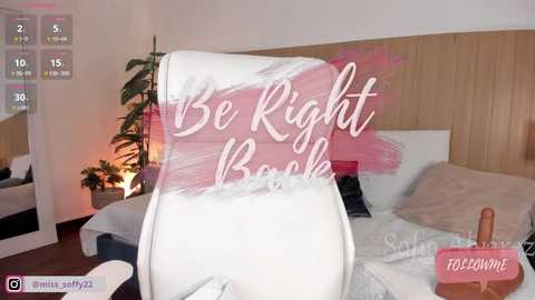 Video of a cozy bedroom with a white duvet and a pink feather boa, captioned \"Be Right Back,\" featuring a man's erect penis in a glass.