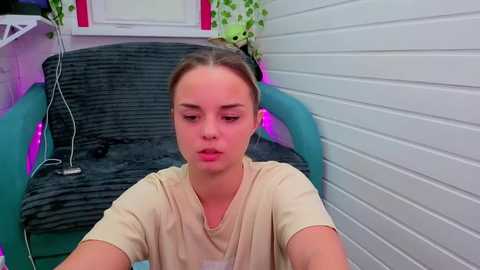Video of a young woman with light skin and brown hair tied back, wearing a beige T-shirt, sitting on a black wicker chair with a green cushion. Background includes a white-paneled wall and a green potted plant with a small stuffed toy.