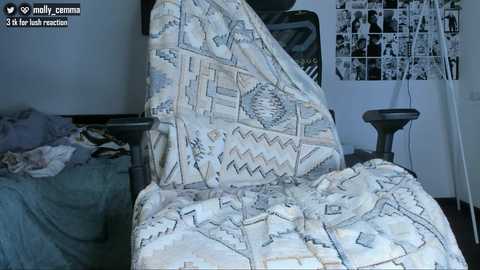A video of a messy bedroom with a beige-patterned quilt covering a black gaming chair, a bed with unmade sheets, and a white wall adorned with a collage of black-and-white photos.