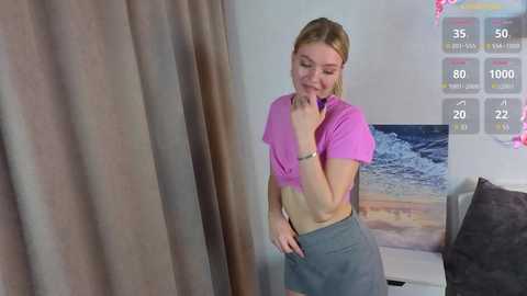 Video of a young, slim, blonde woman with fair skin, wearing a pink crop top and gray skirt, standing near a gray couch in a modern room with a beach mural and digital score display.