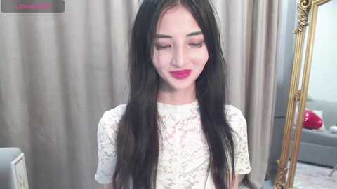 Video of a young Asian woman with long, straight black hair and fair skin, wearing a white lace top, smiling. Background features beige curtains and a gold-framed mirror.