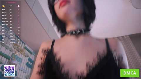 A video of a woman with short black hair, wearing a black lace top and choker, indoors with tiled walls, blurred in the background.