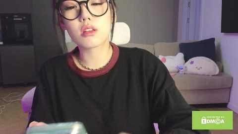 Video of a young Asian woman with glasses and black hair, wearing a black shirt, holding a blue device, in a cozy living room with a beige couch and plush toys.