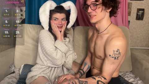 Video of a shirtless, tattooed man with glasses and a woman in a bunny costume on a couch.