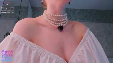 Video of a fair-skinned woman with blonde hair, wearing a white off-shoulder dress, multiple pearl necklaces, and a black pendant, indoors with a tiled wall background.