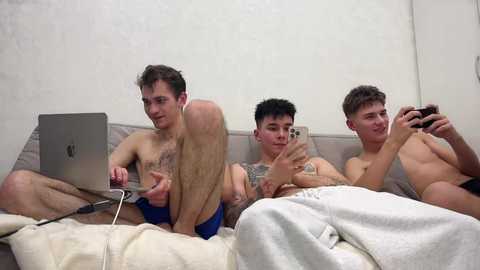 Video of four shirtless young men, two playing video games on a couch, one with a laptop, another in white pants.