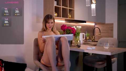 Media: Video of a young woman with light skin, long brown hair, sitting in a modern kitchen, reading a newspaper, wearing lingerie, with a digital display showing weather data.