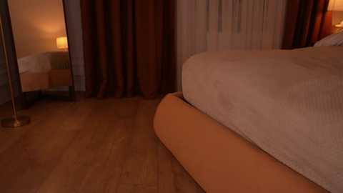 Video of a dimly lit, cozy bedroom featuring a bed with a beige duvet, wooden floor, and maroon curtains.