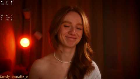 Video of a smiling woman with fair skin and long, wavy red hair, wearing a pearl necklace, against a dimly lit, warm-toned background.