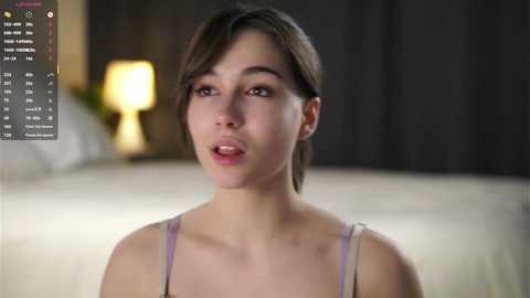 Video of a young Asian woman with short brown hair, wearing a light purple spaghetti strap top, standing in a softly lit bedroom with a yellow lamp.