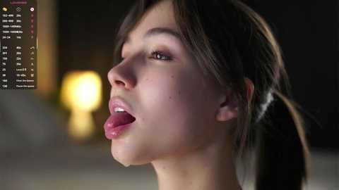 A close-up video of a young woman with fair skin and dark hair, tongue out, in a dimly lit room.