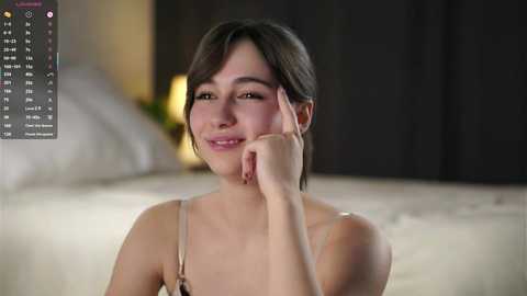 Video of a young Asian woman with short black hair, smiling, resting her chin on her hand, wearing a beige bra, in a dimly lit bedroom.