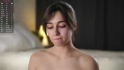 A close-up video of a topless young woman with fair skin, brown hair, and a contemplative expression, taken indoors with a blurred background.