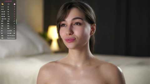 Video of a young, fair-skinned woman with short brown hair, smiling softly, topless, with a blurred background of a dimly lit room featuring a lamp.