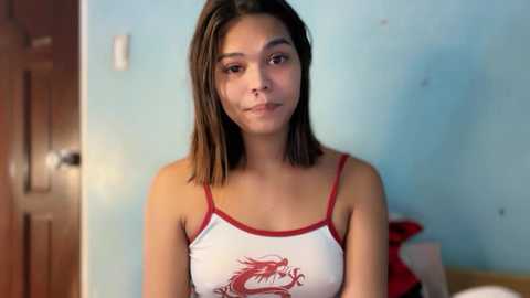 Video of a young Hispanic woman with medium skin tone, shoulder-length dark hair, and a slender build, wearing a white tank top with red trim and a dragon graphic, in a simple, light blue-walled room.