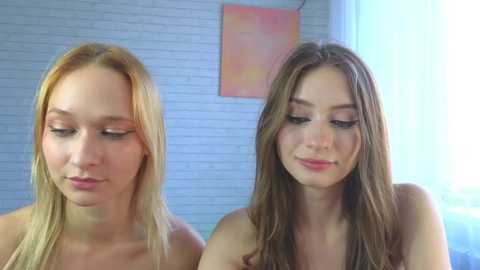 Video of two young Caucasian women with light skin and straight, long hair, one blonde, one brunette, standing side-by-side, topless, in a brightly lit room with a white brick wall and abstract artwork.