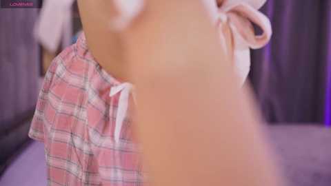 A video of a person wearing plaid pajama shorts, blurred background with purple tones.