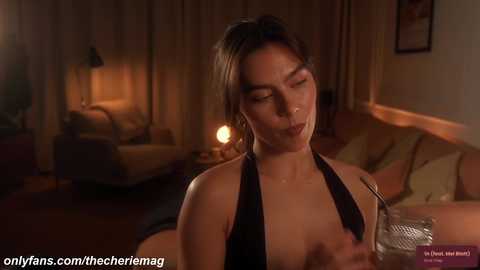 A video of a woman with short dark hair, wearing a black halter top, making a kissy face while holding a glass of water. Background features dimly lit room with a couch and a lamp.