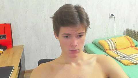 Video of a young, shirtless man with short, dark hair, sitting in a simple room with a bed, a wooden desk, and a red box.