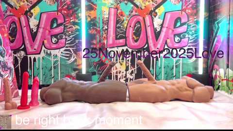 A video of two nude, muscular men, one light-skinned and the other dark-skinned, lying on a bed in a vibrant, graffiti-covered room.