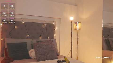 Video of a cozy, well-lit bedroom with a modern, minimalistic design. The bed features a gray headboard, adorned with a string of warm, yellow fairy lights. The room has a soft, ambient glow, creating a serene atmosphere.
