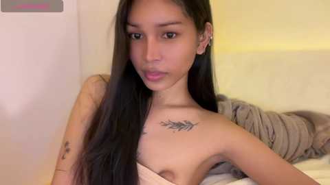 Video of a young, petite Asian woman with long black hair, bare-chested, showcasing a small, detailed feather tattoo on her chest, lying on a beige bed.