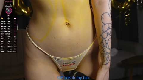 Video of a light-skinned woman with tattoos, wearing a small, beige thong with \"woofer\" printed on it, indoors, blurred background.