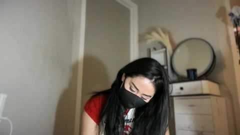 Video of a young woman with long black hair, wearing a red t-shirt, black face mask, and dark glasses, leaning over a wooden dresser with a round mirror, in a dimly lit room with beige walls.
