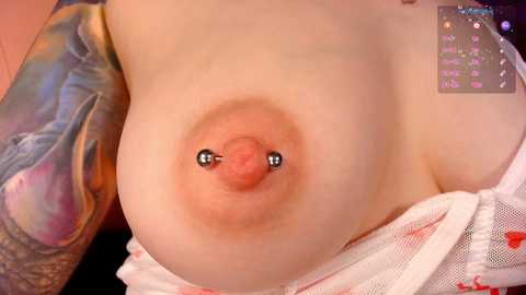 A close-up video of a fair-skinned woman's bare breast with a silver nipple piercing and an elaborate tattoo sleeve. She wears a white lace garment partially pulled down.