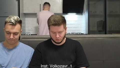 Video of two men in a modern kitchen: one with short blonde hair, blue shirt, and another with short brown hair, black shirt. Background features a white-tiled wall and stainless steel appliances.