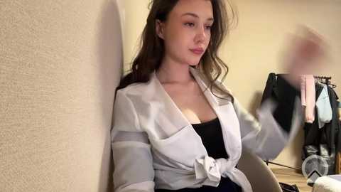 Video of a young woman with long, wavy brown hair, wearing a white robe over a black top, sitting against a beige wall. Background features a clothing rack with various garments.