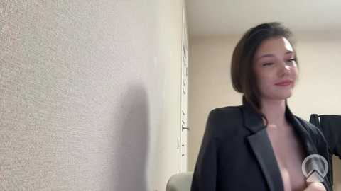 Video of a young Asian woman with short black hair, wearing a black blazer, standing in an office corridor, holding a microphone, smiling slightly.