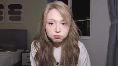 Video of a young woman with long, blonde hair and pale skin, wearing a white shirt, making a pouty face in a dimly-lit bedroom with a bed and abstract artwork on the wall.