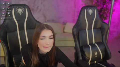 Video of a young woman with long brown hair, fair skin, wearing black clothing, smiling, kneeling on a black gaming chair with white stripes in a dimly lit room with purple lighting.