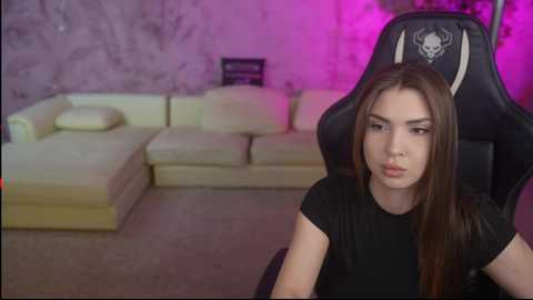 A video of a young woman with long brown hair, wearing a black T-shirt, sitting in a black gaming chair with a skull emblem, in a dimly-lit living room with a beige sectional sofa.
