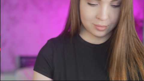 Video of a young woman with long, straight, light brown hair, wearing a black T-shirt, and standing in a softly lit room with purple-pink lighting.
