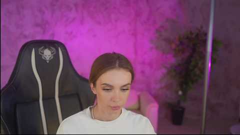Video of a young woman with light skin and brown hair, sitting in a black gaming chair with a skull logo, wearing a white shirt, in a dimly lit room with pink and purple lighting.